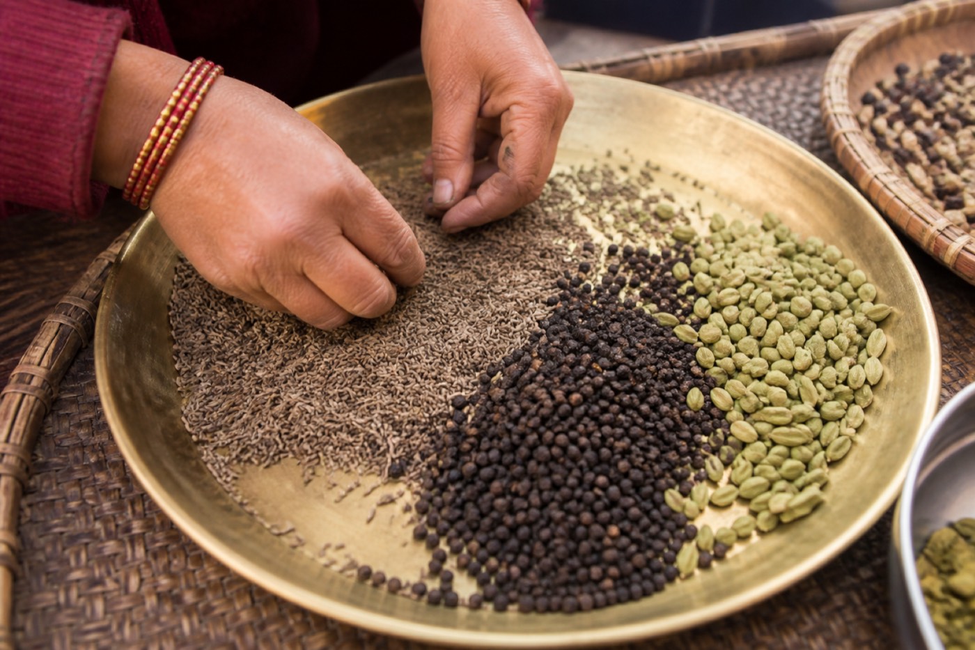 Hands sorting whole cumin, pepper and cardamom on a brass tray